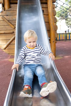 A little blonde boy slide down a slide on the playground