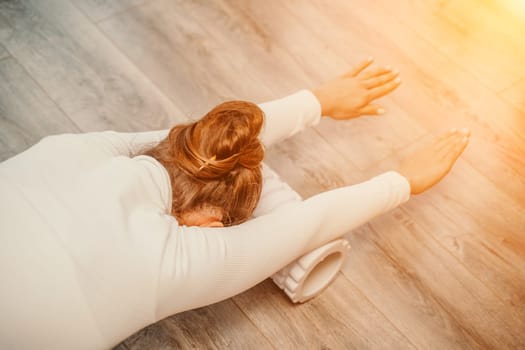 Adult athletic woman, in white bodysuit, performing fascia exercises on the floor - caucasian woman using a massage foam roller - a tool to relieve tension in the back and relieve muscle pain - the concept of physiotherapy and stretching training