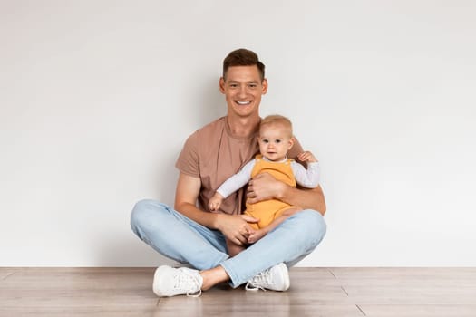 Happy Young Dad With Infant Son Sitting On Floor Near White Wall