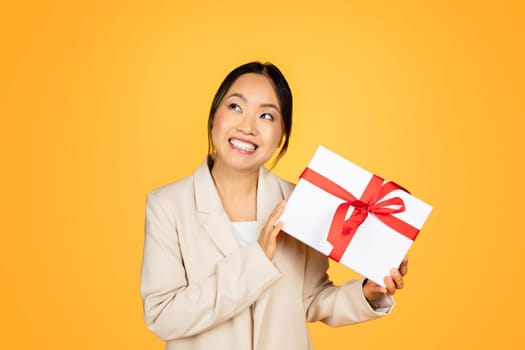 Smiling Asian millennial woman joyfully holding a wrapped holiday gift, festive atmosphere around