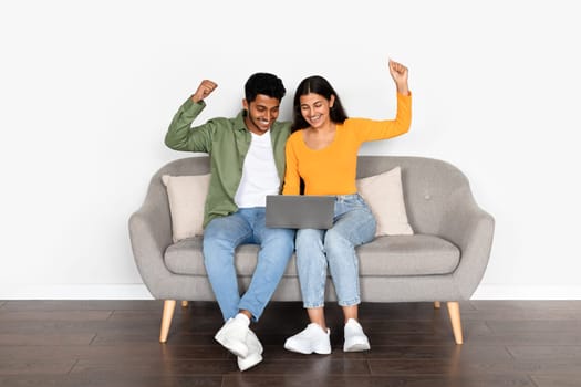 Excited indian couple shaking fists over laptop sitting on sofa