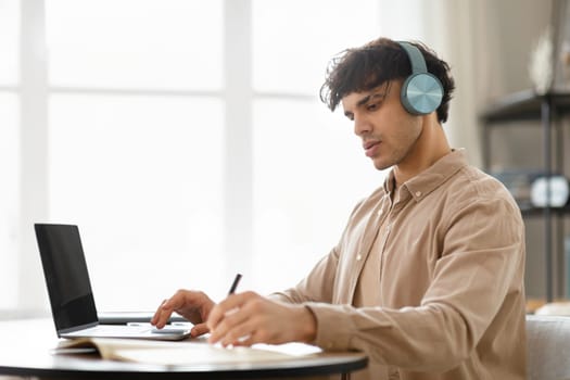 Arab Man At Laptop Writing Notes Wearing Wireless Earphones Indoors