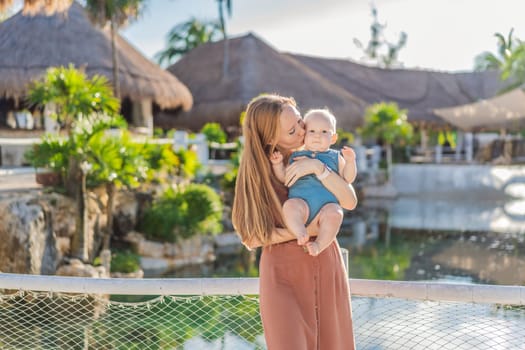 Amidst tropical palms and thatched roofs, a loving mom embraces her baby, sharing warmth and affection in a tranquil outdoor setting