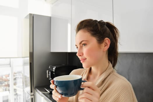 Close up portrait of beautiful young woman, posing in the kitchen, looking dreamy and thoughtful, contemplating morning with mug of coffee, waiting for breakfast.