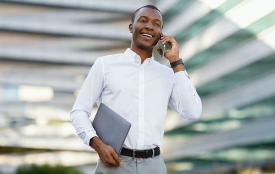 Business Call. Smiling Black Businessman Talking On Cellphone While Walking Outdoors Near Modern Office Center, Handsome African American Entrepreneur Holding Laptop, Enjoying Pleasant Conversation