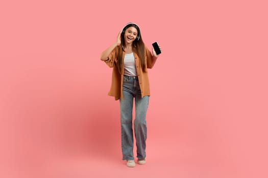 Positive teen girl in wireless headphones listening music on smartphone and dancing