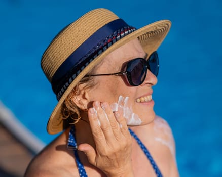 Portrait of an old woman in a straw hat, sunglasses and a swimsuit applying sunscreen to her face while relaxing by the pool.