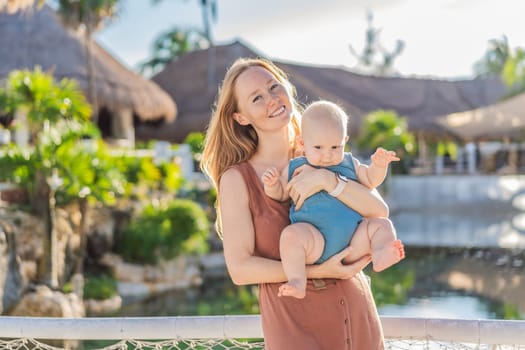 Amidst tropical palms and thatched roofs, a loving mom embraces her baby, sharing warmth and affection in a tranquil outdoor setting