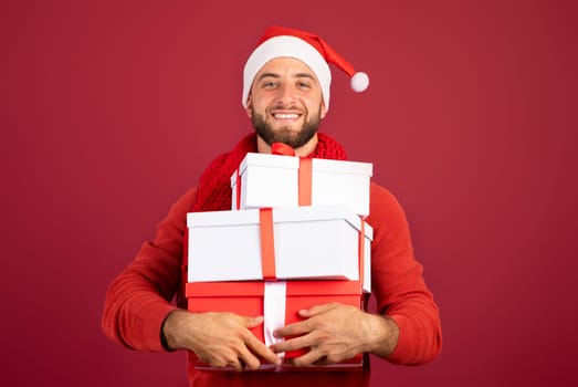 Cheerful handsome millennial caucasian man in Santa hat hold boxes of gifts, enjoy holidays