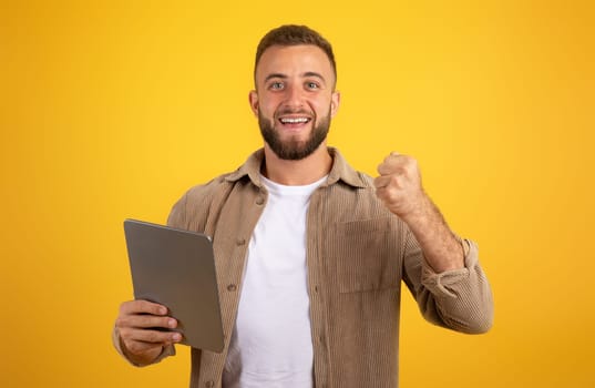 Cheerful confident handsome european millennial man with beard use tablet, celebrate win with fist sign