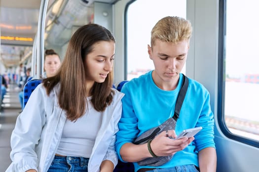 Teenage guy and girl commuter train passengers sitting together looking at smartphone
