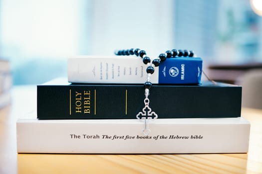 Books, stack and closeup on table for faith, Abrahamic religion or rosary with crucifix for study in home. Knowledge, holy spirit and education with cross for Jesus, Muhammad or Moses with solidarity.