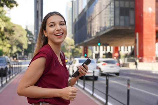 Portrait of young Brazilian woman holding mobile phone looking at camera on Paulista Avenue with blurred background in Sao Paulo, Brazil