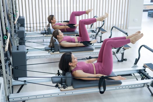 Three Asian women in pink sportswear doing pilates exercises with a reformer bed.