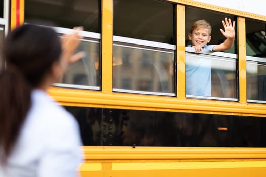 Boy Sitting Inside Of Yellow School Bus And Waving Hand To Mother