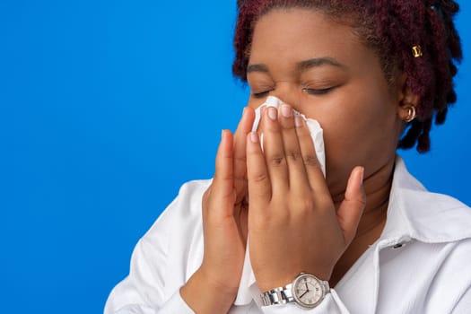 Sick afro woman sneezing in paper napkin against blue background