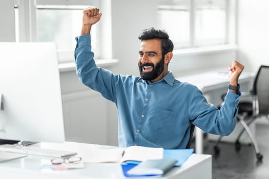 Indian businessman looking at computer monitor and shaking fists celebrating big luck in office
