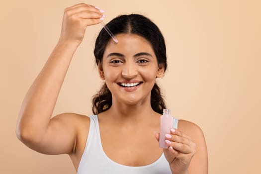 Beauty Routine Concept. Beautiful Indian Female Applying Face Serum With Dropper While Posing Over Beige Studio Background, Smiling Eastern Woman Moisturizing Skin, Enjoying New Beauty Product