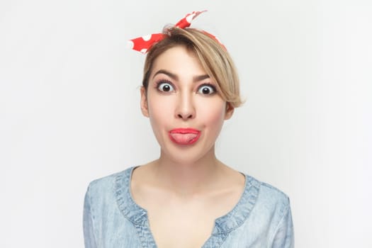 Funny foolish woman in denim shirt and red headband standing sticking tongue out, looking at camera.