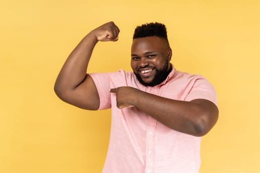 Portrait of smiling man wearing pink shirt pointing finger at arm biceps, showing his strength and independent, leadership concept. Indoor studio shot isolated on yellow background.