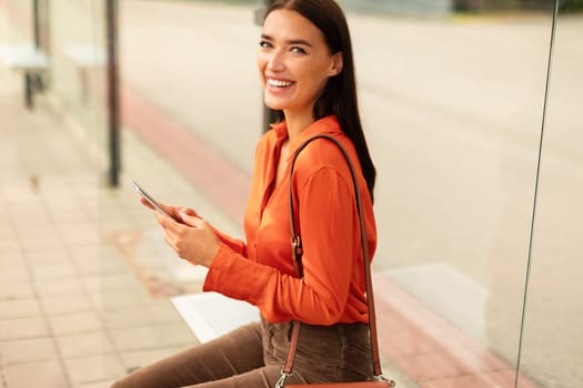 Cheerful passenger woman holds phone booking tram tickets at stop