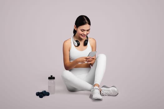 Fit woman with headphones, checking phone post-exercise on lavender background