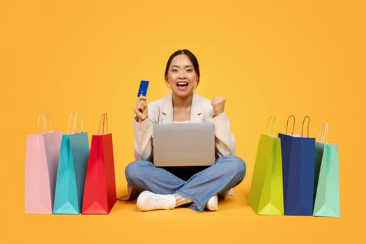 Glad excited millennial chinese lady with shopping bags, credit card, laptop, enjoy sale and win
