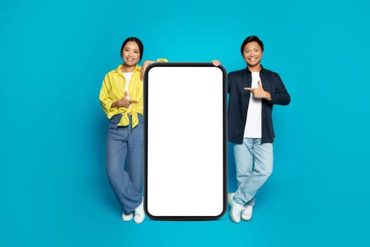 Cheerful asian couple in casual pointing fingers at large smartphone with empty space