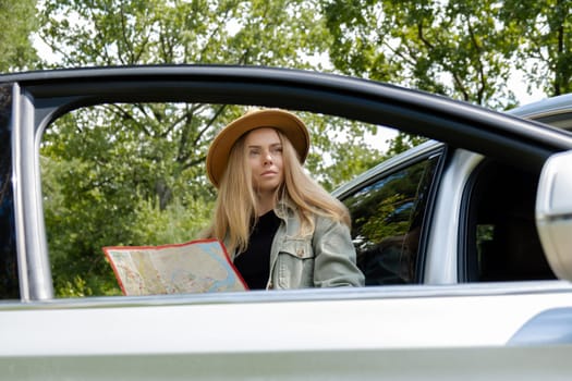 Smiling young woman in hat stoped on road to check the route on travel map. Local solo travel on weekends concept. Exited woman explore freedom outdoors in forest. Unity with nature lifestyle, rest recharge