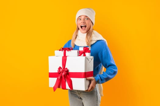 Joyful woman holding Christmas boxes in a yellow studio, exuding excitement and festive spirit