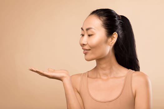 Serene middle-aged Asian woman, extending her hand and presenting new beauty product on her palm, shot in a studio with a soft beige backdrop