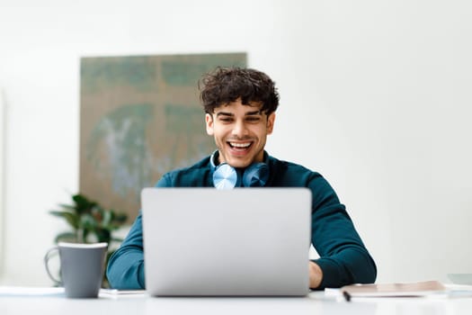 Happy european freelancer man working on laptop wearing headphones on neck and smiling, sitting in light office