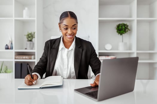 Cheerful black woman entrepreneur attending online business meeting, lady sitting in front of laptop and taking notes