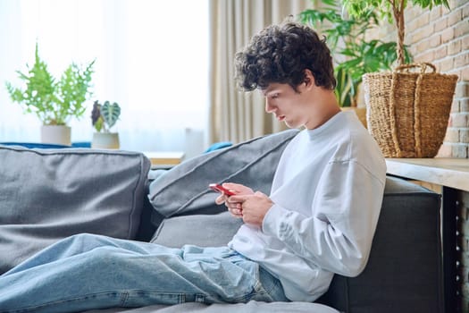 Young handsome guy using smartphone sitting on sofa in living room. Male 20 years old with curly fashionable hairstyle. Internet online technology mobile applications for study leisure communication