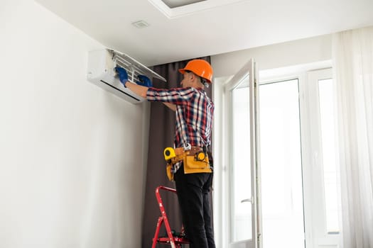 Worker installing or repairing air conditioner. Young man in uniform standing on ladder with toolbox and installing new air conditioner on wall. AC installation, maintenance and repair service concept