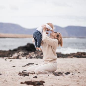 Mother enjoying winter vacations holding, playing and lifting his infant baby boy son high in the air on sandy beach on Lanzarote island, Spain. Family travel and vacations concept