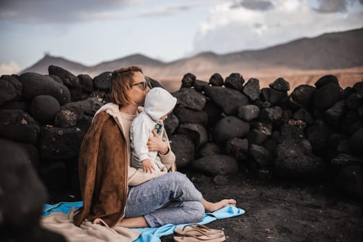 Mother enjoying winter vacations playing with his infant baby boy son on black sandy volcanic beach of Janubio on Lanzarote island, Spain on windy overcast day. Family travel vacations concept.