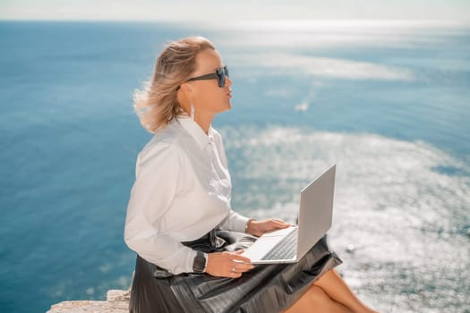 Business woman on nature in white shirt and black skirt. She works with an iPad in the open air with a beautiful view of the sea. The concept of remote work.