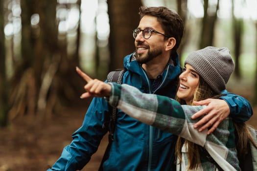 Glad young caucasian couple in jackets enjoy journey, points finger at copy space, rest in forest