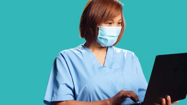 Close up of smiling happy healthcare specialist using laptop to look up medical informations online. Asian nurse wearing professional clinical protective uniform, isolated over studio background
