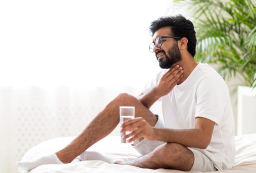 Sick indian man suffering throat ache while sitting on bed at home