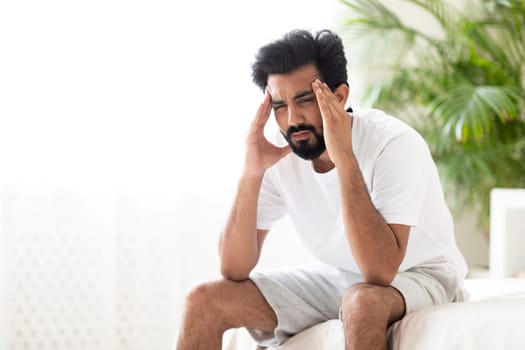 Portrait Of Depressed Young Indian Man Sitting On Bed At Home