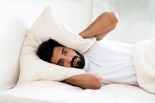 Sleepless young indian man lying in bed and covering ears with pillow