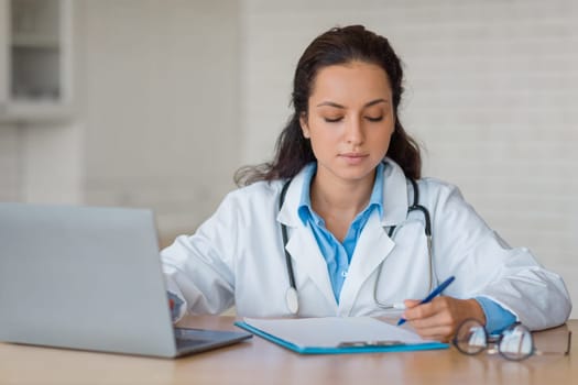 Focused doctor writing notes with laptop nearby
