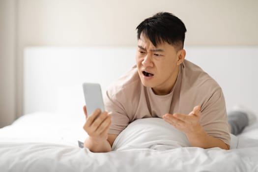 Angry middle aged asian man lying on bed with phone