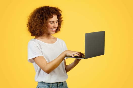 Glad young european lady student typing on laptop, chatting in social networks, isolated on yellow background, studio. Technology for work, productivity, business remotely and modern app