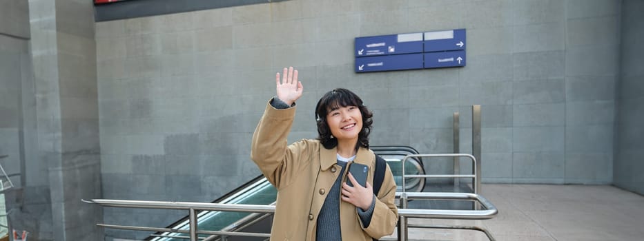 Portrait of cute smiling asian girl in headphones, stands on street with smartphone, waves hand at someone, says hello, greets friend.