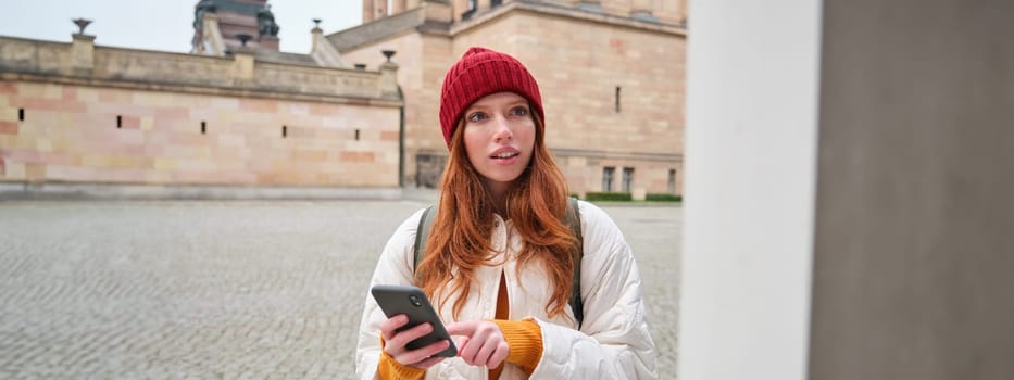 Tourism and sightseeing concept. Young redhead woman, tourist walks around city, looks at her smartphone app and at history stand, explores adventures.
