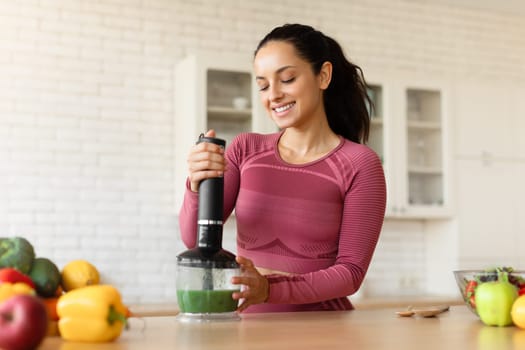Smiling woman in fitwear making smoothie with blender in kitchen