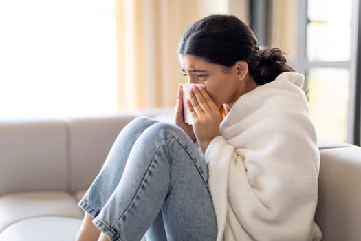 Sick Young Indian Woman Blowing Nose In Tissue While Sitting On Couch